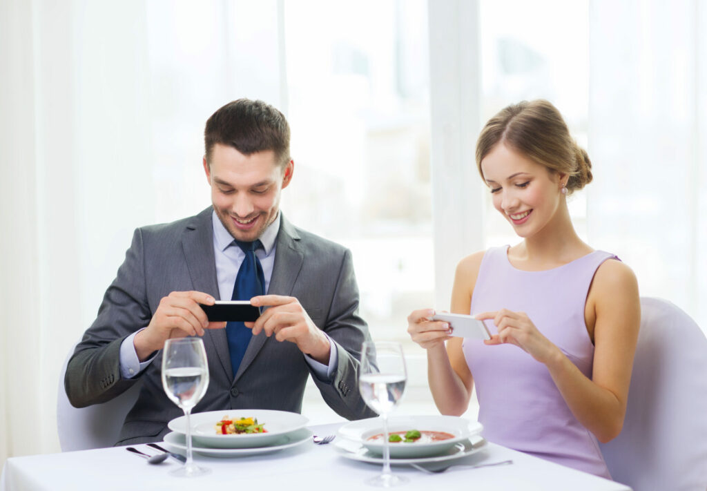 Well dressed couple in nice restaurant taking photos on their phones ready to send to WhatsApp chat groups 