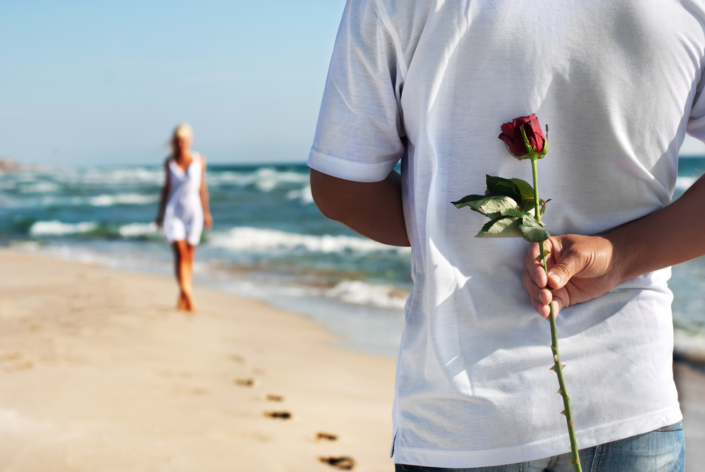 man with red rose behind his back standing on beach with his love coming towards him 