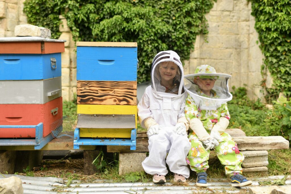 kids sat next to a beehive in beekeeping attire
