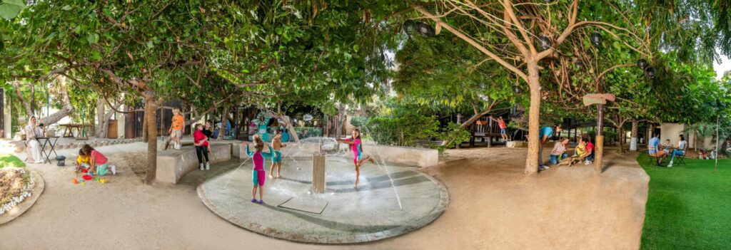 a green play space with trees and kids running in a splash pad 