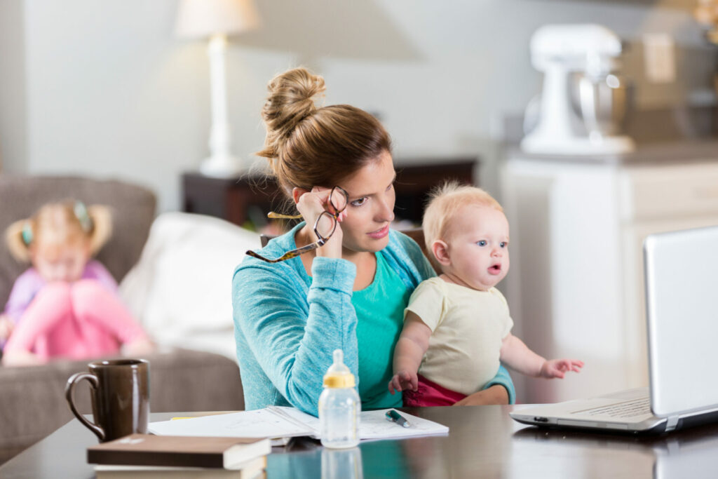 Concerned mom with mum guilt uses a laptop while working. She is holding her baby girl. 