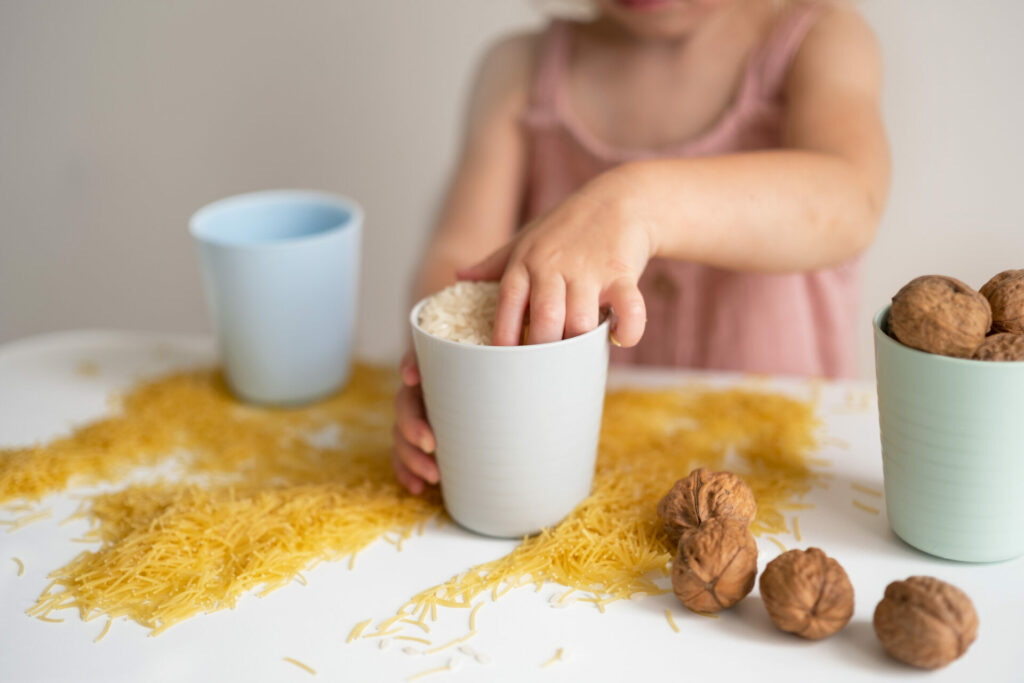 Baby girl, toddler playing with grain, nuts, pasta and rice at table.Sensorial early development, education for baby, child.Kid useful games.