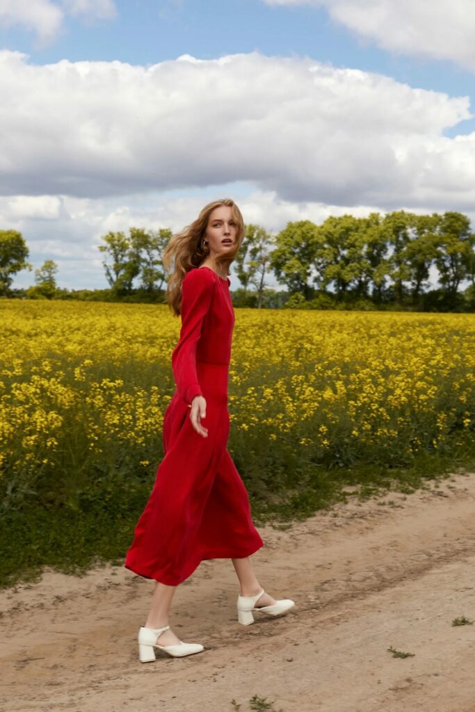 model in red dress walks near yellow rapeseed oil field 