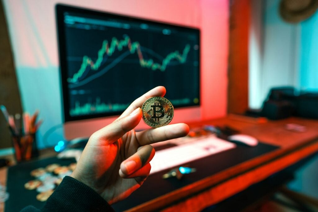 a hand holding a crypto coin in front of a computer screen