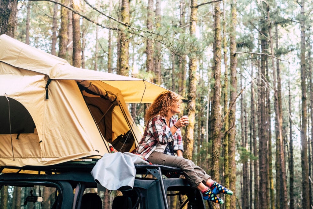 lady sits on tent on top of van in the woods drinking coffee from cup