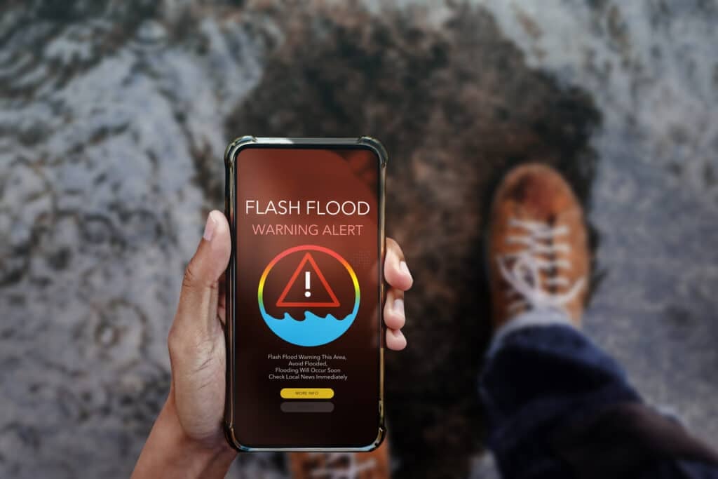 Smartphone displaying a flash flood warning alert screen, held by a person standing near rising floodwaters. Concept for emergency notification, natural disaster preparedness, and real-time weather alerts
