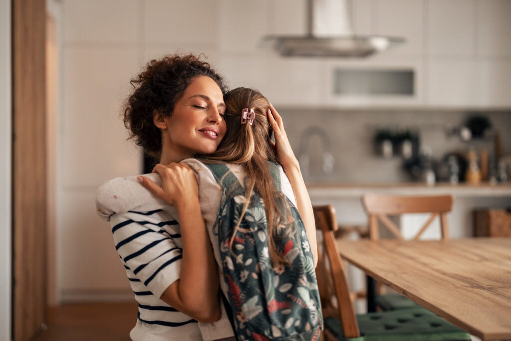 A heartwarming moment of a mother embracing her young daughter, who is wearing a backpack, in a cozy kitchen. The image conveys love, family connections, teaching empathy and preparation for school.