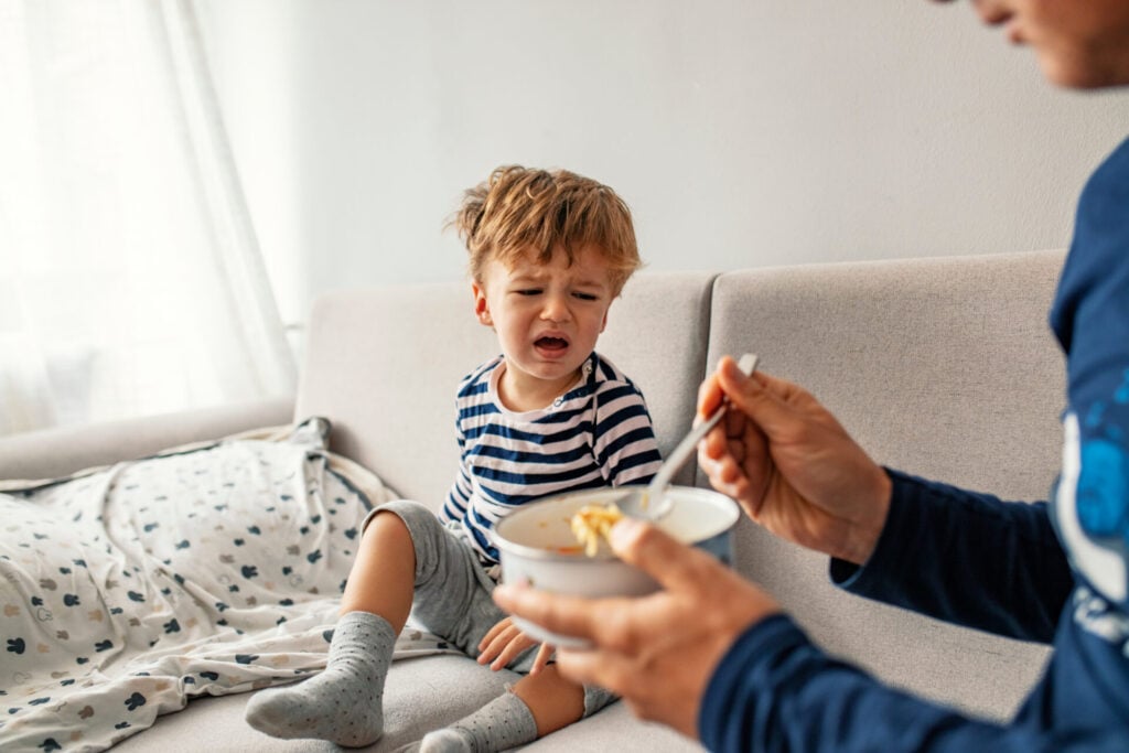 little boy crying with father trying to make him eat some pasta from a bowl 