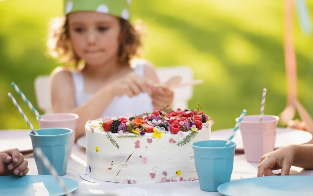 little girl with hat on and homemade birthday cake in front of her with edible flower and berries 