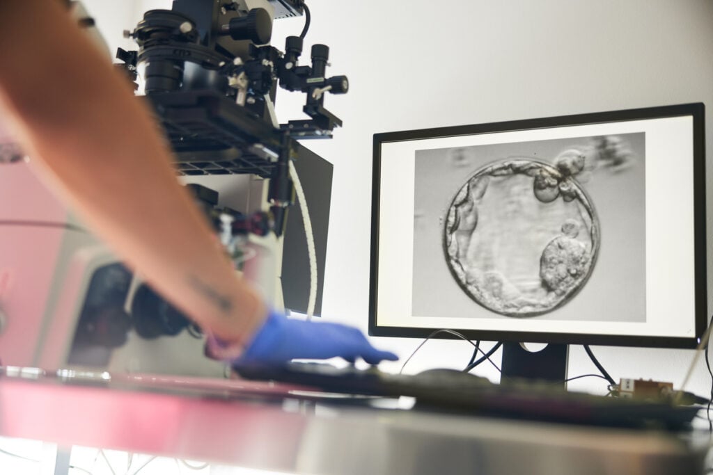 A fertility procedure is shown on the screen while a woman in scrubs follows the process in the lab.