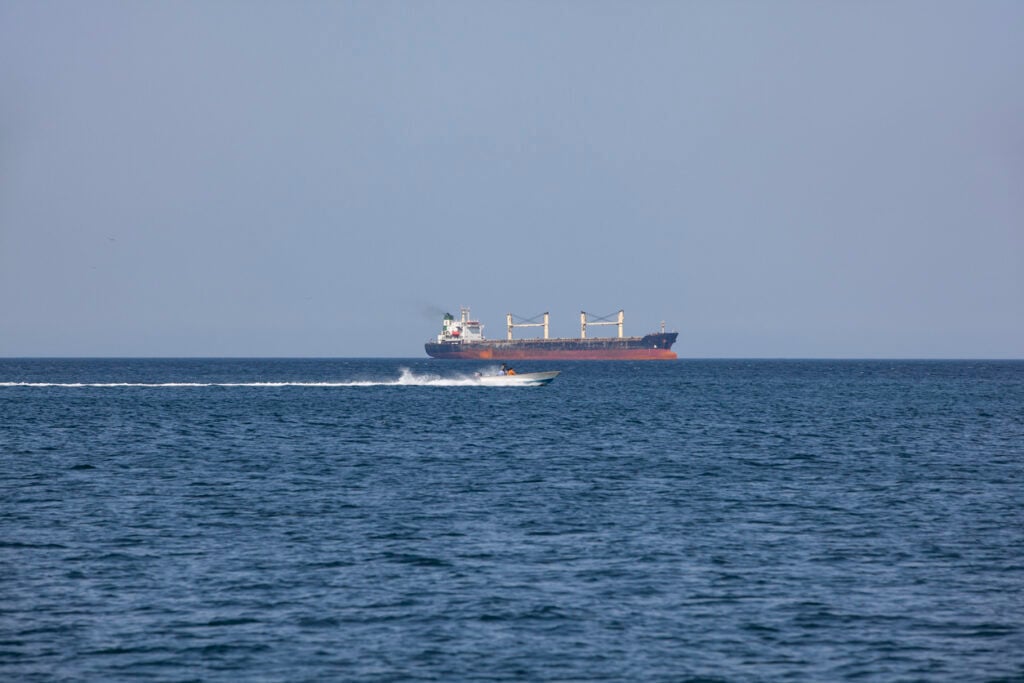 Large cargo vessel sailing in the Strait of Hormuz with a small speedboat passing in the foreground. The strategic waterway between Oman and Iran is one of the world’s most important maritime shipping routes.