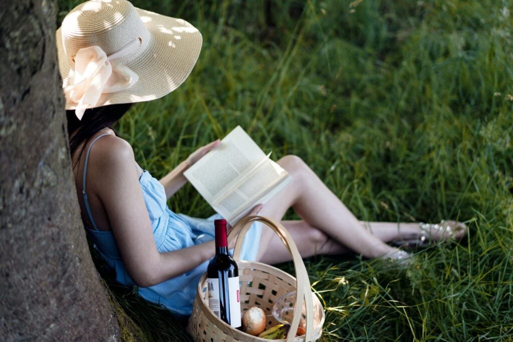 woman reading under the shade of trees with picnic in basket next to her 