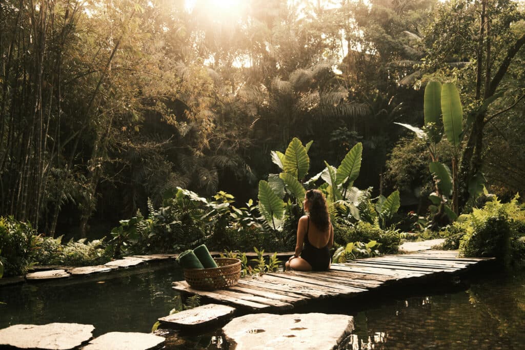 Woman sitting on a wooden pier by a natural pond, finding tranquility and wellness in a lush tropical resort