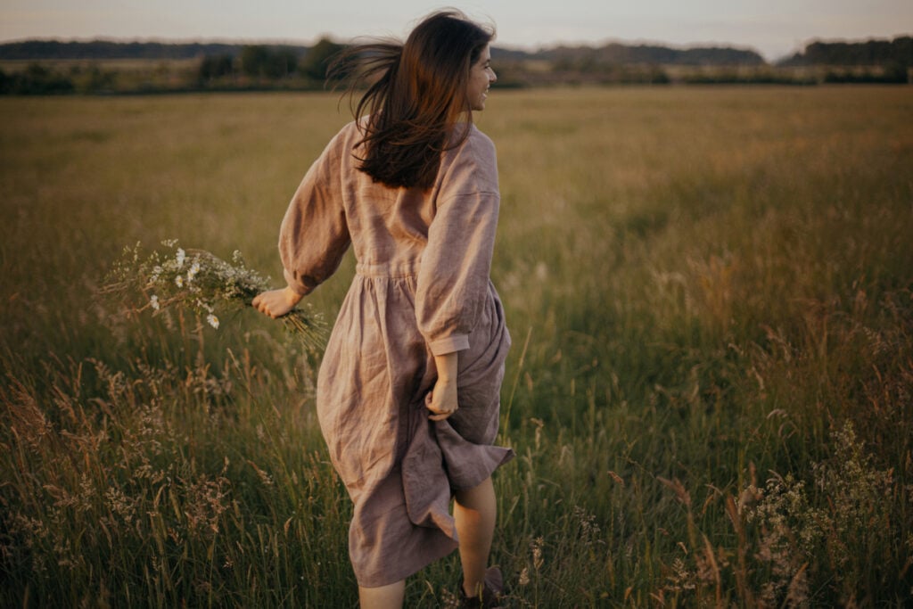 Beautiful woman in linen dress running with wildflowers in hand in summer meadow in sunset. Atmospheric carefree moment. Stylish young female in rustic dress enjoying free evening in countryside