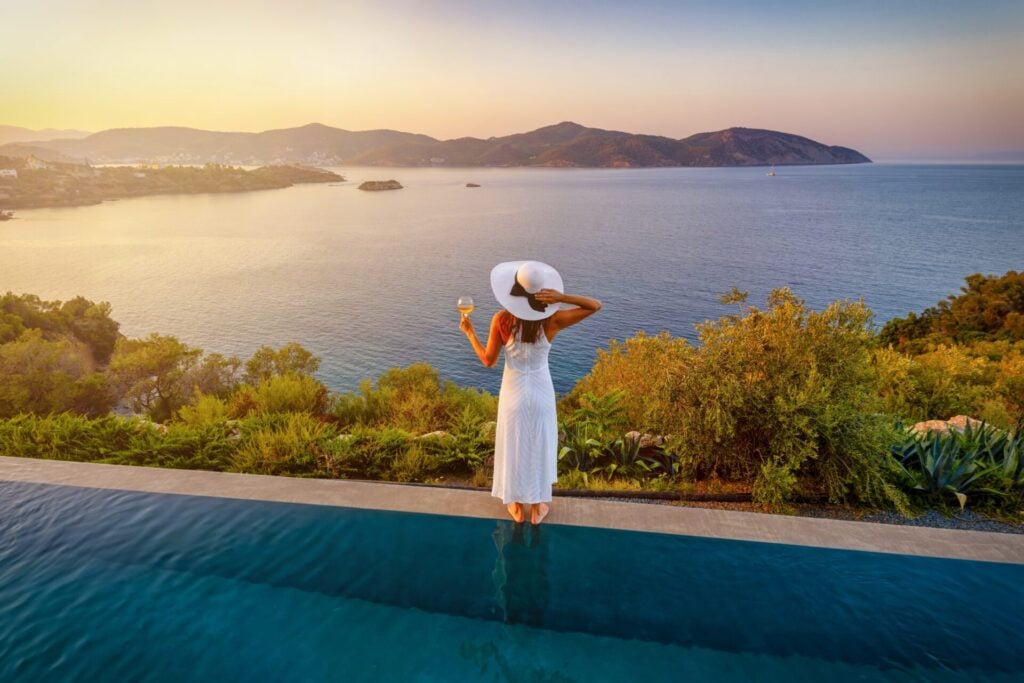 Woman looking out to sea in a white dress and summer hat holding a glass of wine 