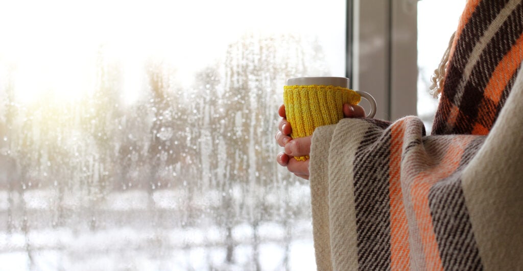 Woman in a blanket with a mug in hands against the background of a window with raindrops on a cold day